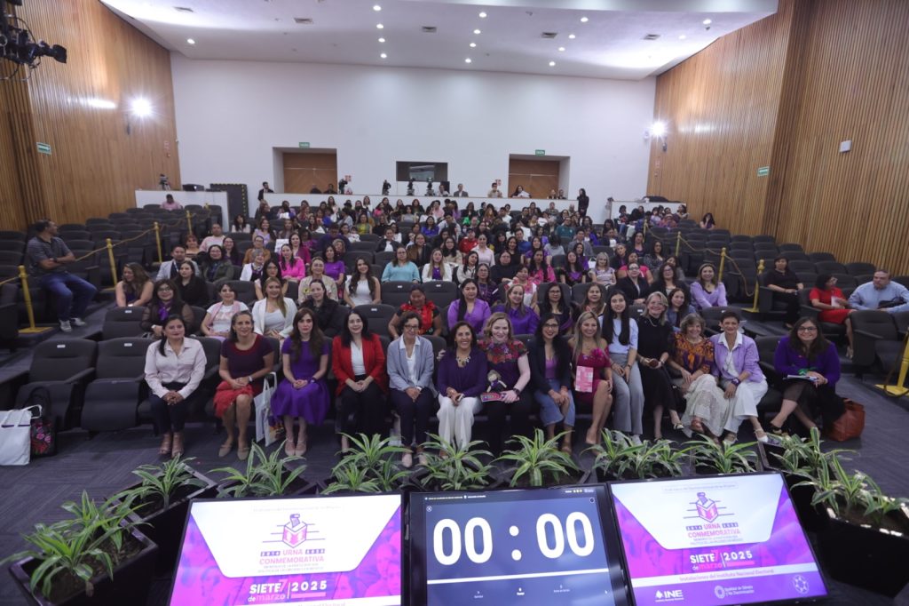 Imagen abierta en el auditorio del instituto con las personas participantes paradas en el estrado al frente del recinto y letrero del Instituto Nacional Electoral