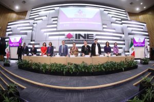 Imagen abierta en el auditorio del instituto con las personas participantes de pie al fondo del recinto, pantalla con logo del evento contiene una urna y abajo de ella letrero del Instituto Nacional Electoral.