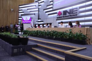 Imagen abierta en el auditorio del instituto con las personas participantes sentadas en el estrado al frente del recinto, pantalla con logo del evento contiene una urna y abajo de ella letrero del Instituto Nacional Electoral.