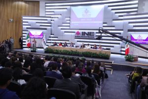 Imagen abierta en el auditorio del instituto con las personas participantes sentadas en el estrado al frente del recinto, pantalla con logo del evento contiene una urna y abajo de ella letrero del Instituto Nacional Electoral.