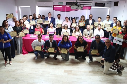 Varias personas de pie y otras hincadas sosteniendo un reconocimiento
