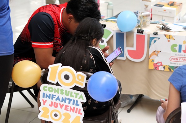 Una menor sentada auxiliada por un adulto en la Consulta Infantil y Juvenil 2024