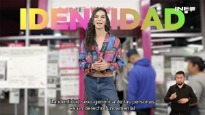 Persona Trans de pie con un letrero a sus espaldas que dice "Identidad" con un módulo de atención ciudadana al fondo