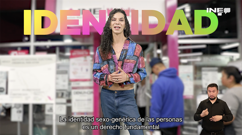 Persona Trans de pie con un letrero a sus espaldas que dice "Identidad" con un módulo de atención ciudadana al fondo