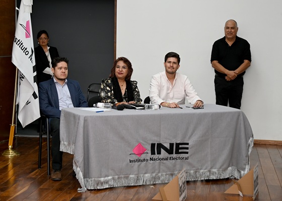 tres personas sentadas frente a una mesa con un mantel gris y la leyenda INE
