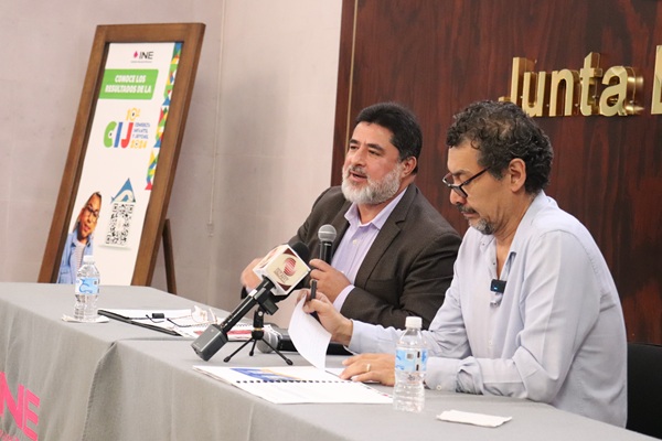 dos hombres sentados, uno habla mediante un micrófono