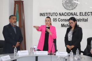 mujer de saco largo rosa con micrófono en una mano y la otra levantada hacia el frente