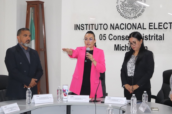 mujer de saco largo rosa con micrófono en una mano y la otra levantada hacia el frente en señal de juramento