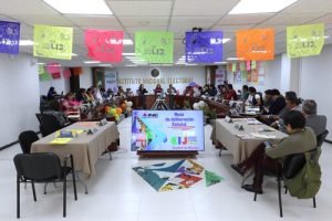 personas sentadas frente en una mesa ovalada y en medio una pantalla