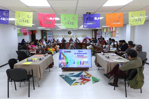 personas sentadas frente en una mesa ovalada y en medio una pantalla