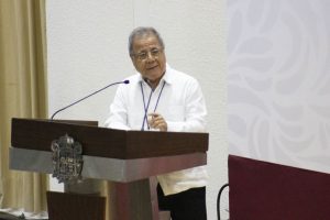Hombre de pie, lentes, camisa blanca hablando en un micrófono