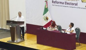 un adulto mayor de pie frente a un atril, a su lado dos mujeres sentadas