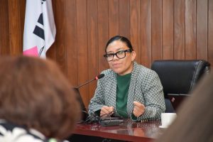 un mujer sentada, cabello recogido hablando frente a un micrófono