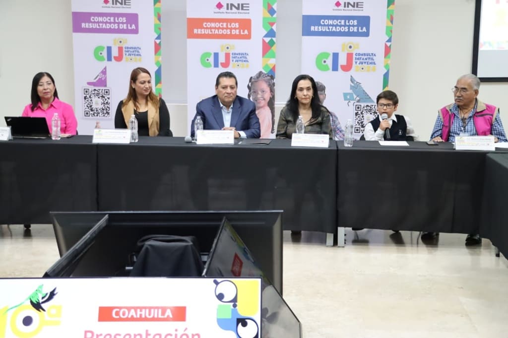 mujeres, hombres y un niño sentados frente a una mesa con micrófonos