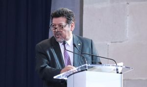 hombre de traje, lentes, de pie frente a un atril con micrófono