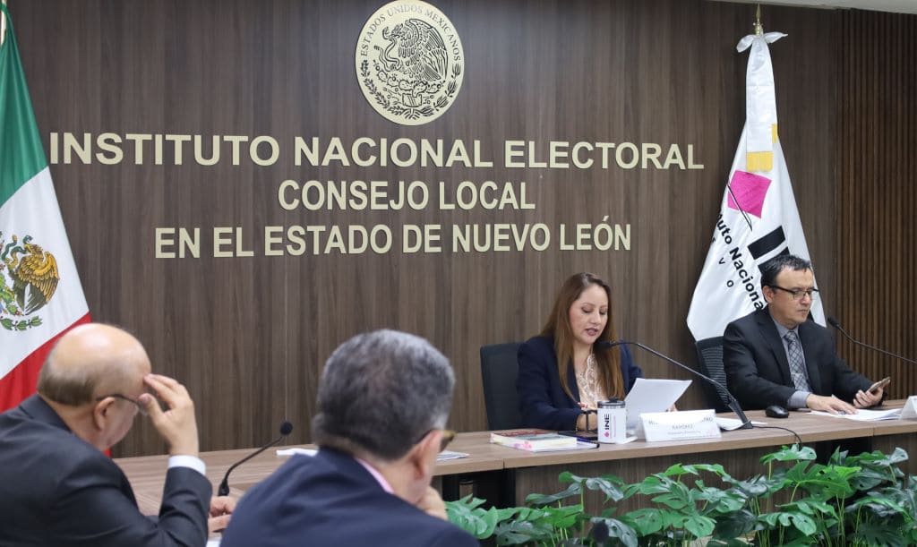 personas sentadas en un salón con bandera de México y del INE