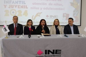 tres mujeres y dos hombre sentados atrás de una mesa con mantel y las letras INE