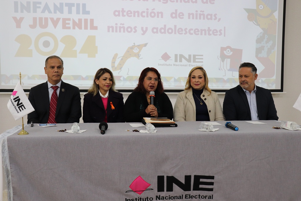 tres mujeres y dos hombre sentados atrás de una mesa con mantel y las letras INE