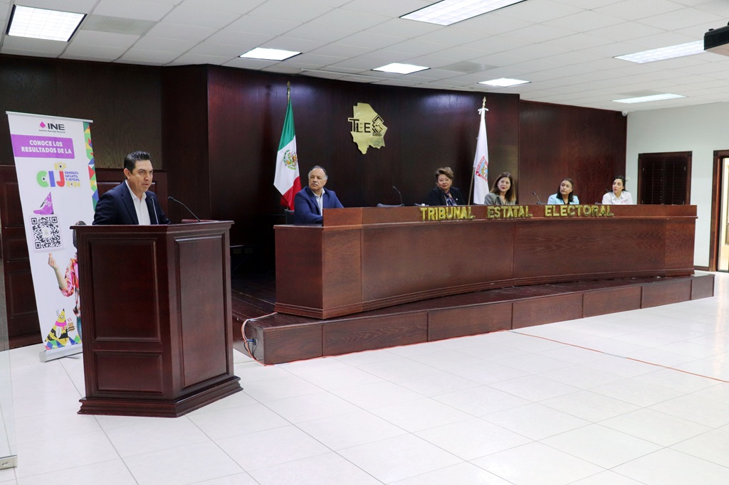mesa grande de madera, personas sentadas atrás y un hombre de pie hablando