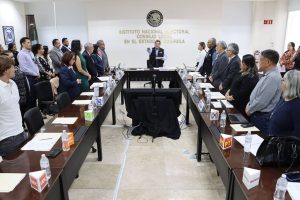 personas de pie al rededor de una mesa rectangular con micrófonos