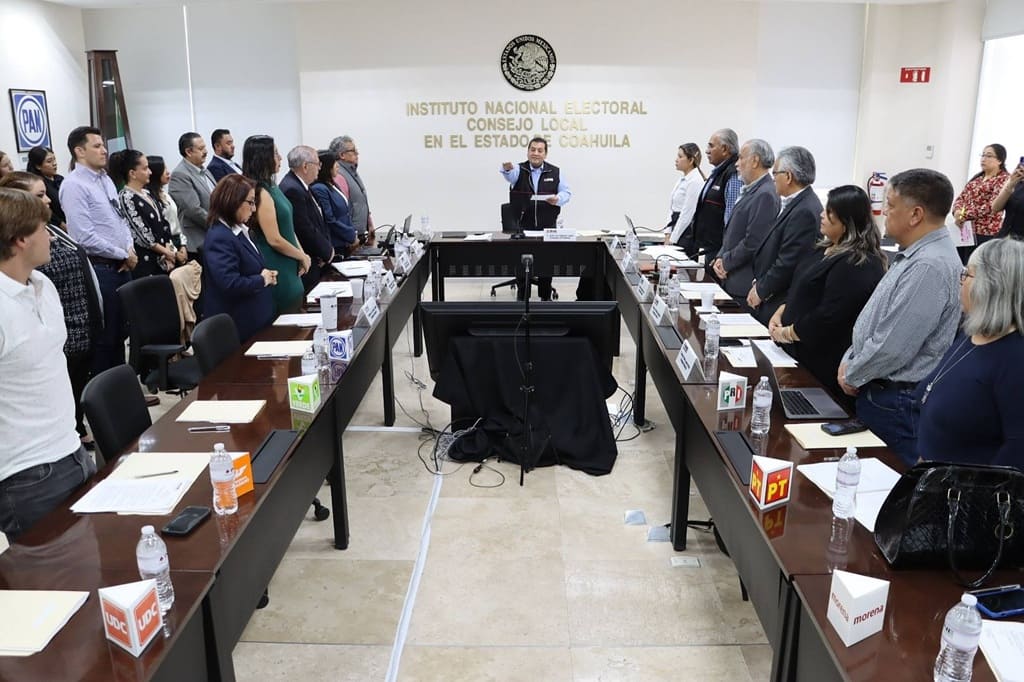 personas de pie al rededor de una mesa rectangular con micrófonos