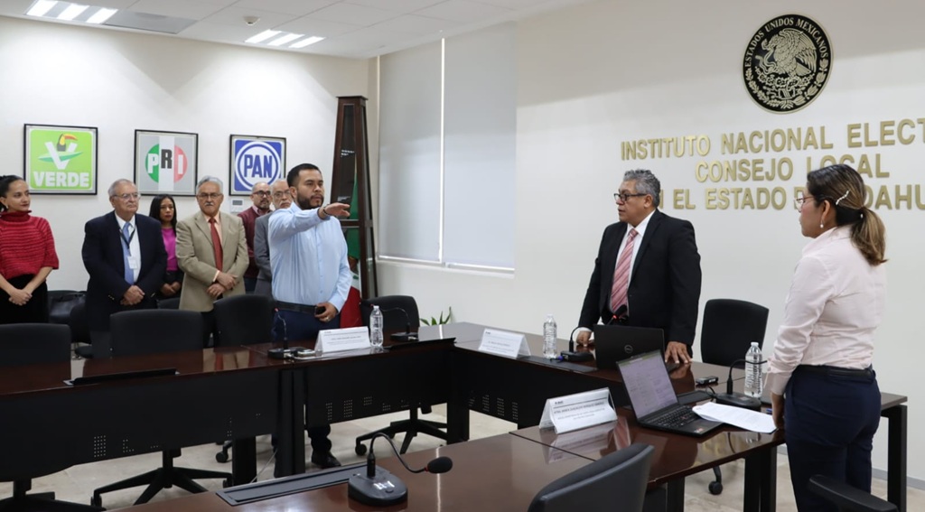 varias personas de pie en un salón, un hombre levanta la mano derecha al frente