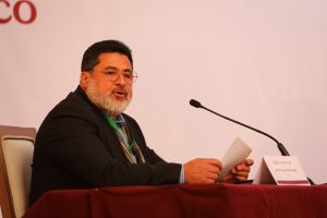 un hombre sentado de barba blanca con lentes hablando frente a un micrófono