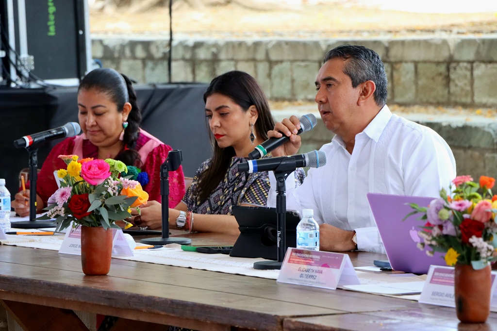 hombre sentado hablando frente a un micrófono, a su lado, dos mujeres