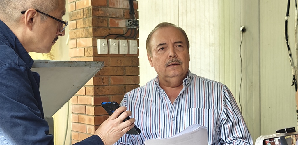 Un hombre de pie sosteniendo un documento hablando frente a persona que sostiene un celular