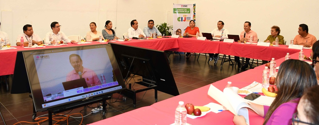 varias personas sentadas alrededor de una mesa con mantel rosa, en medio una pantalla de televisión