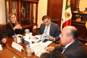 dos hombres y una mujer sentados en una oficina con muebles de madera y una bandera de México