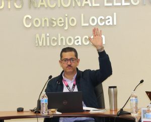 un hombre sentado frente a un micrófono con lentes votando a mano alzada