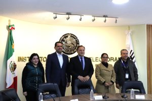 tres mujeres y dos hombre de pie frente al escudo nacional y la bandera de México