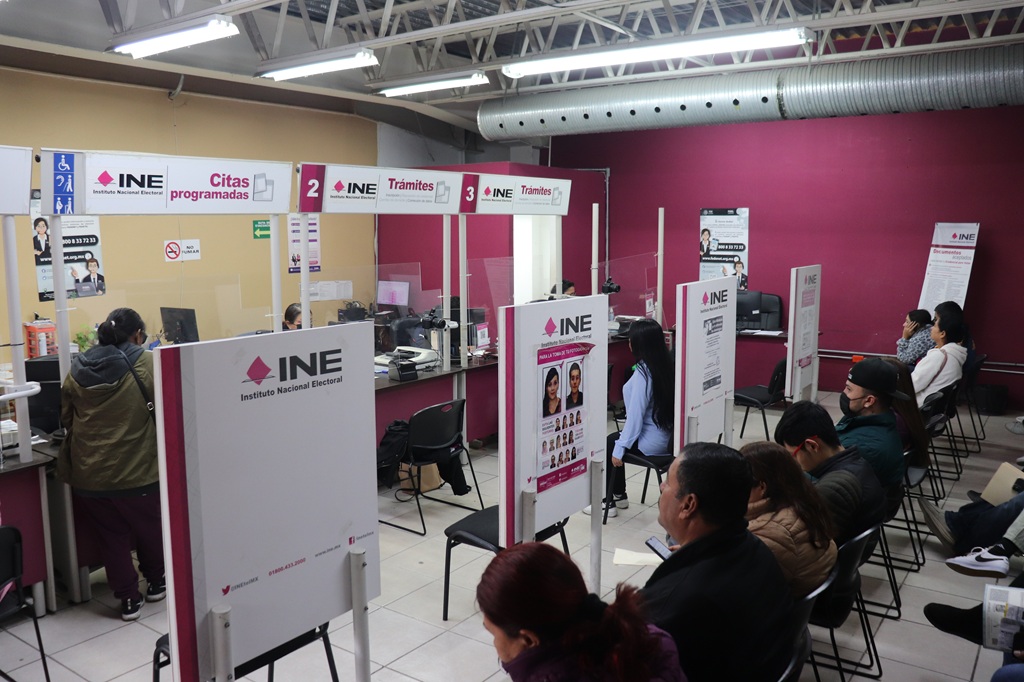 personas sentadas frente a mamparas color rosa y blanco con leyenda INE