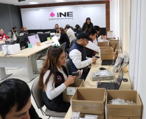 personas con chalecos negros sentados frente a cajas con credenciales para votar sin validez
