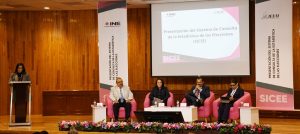 personas sentadas en un escenario, a su lado una persona de pie hablando