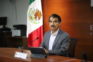 un hombre sentado frente a un micrófono, detrás la bandera de México