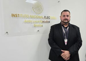 un hombre de barba y saco negro de pie a lado de un letrero