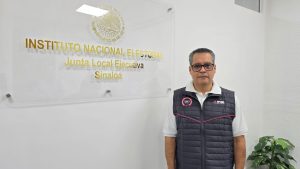 un hombre con chaleco negro y lentes de pie a lado de un letrero