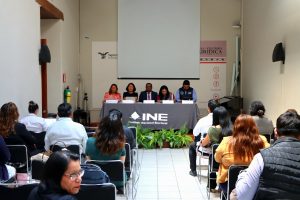 un salón con personas sentadas, viendo a cuatro personas al frente