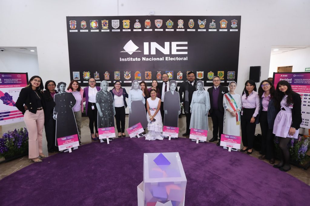 Personas participantes paradas en el lobby del auditorio posando con las fotos de mujeres destacadas en la vida política y una urna.