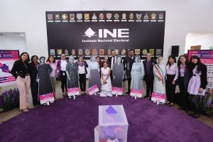 Personas participantes paradas en el lobby del auditorio posando con las fotos de mujeres destacadas en la vida política y una urna.