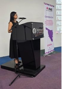 Una mujer de pie con vestido negro de frente a un atril, hablando a través de un micrófono