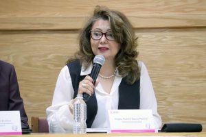 Mujer con cabello largo y lentes, vestida con camisa blanca hablando a través de un micrófono.
