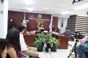 Ocho personas sentadas, reunidas al rededor de una mesa, hablando detrás de micrófonos.
