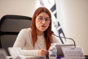 Mujer sentada, cabello rojizo lacio y camisa blanca, habla a través de un micrófono