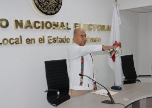 Hombre de pie, calvo, guayabera blanca con el brazo derecho extendido hacia el frente