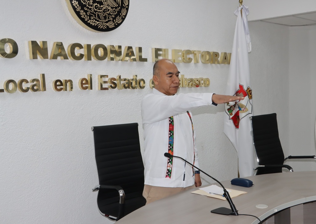 Hombre de pie, calvo, guayabera blanca con el brazo derecho extendido hacia el frente