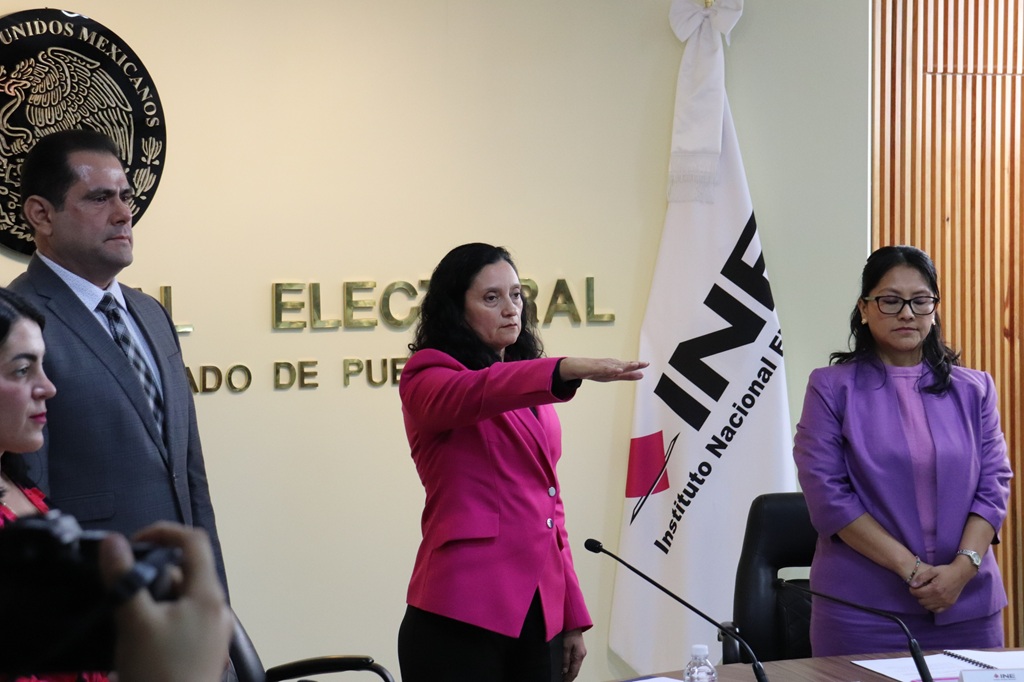 mujer de pie, cabello rizado al hombro, saco rosa, con el brazo derecho al frente