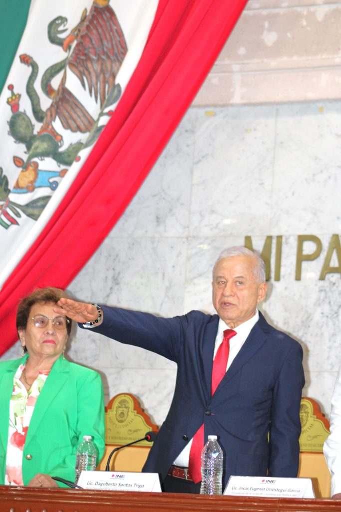 Hombre de pelo cano vestido de traje de pie detrás de una mesa